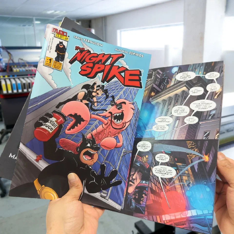 A copy of The Night Spike #1 and an interior spread, held in front of the press that printed them